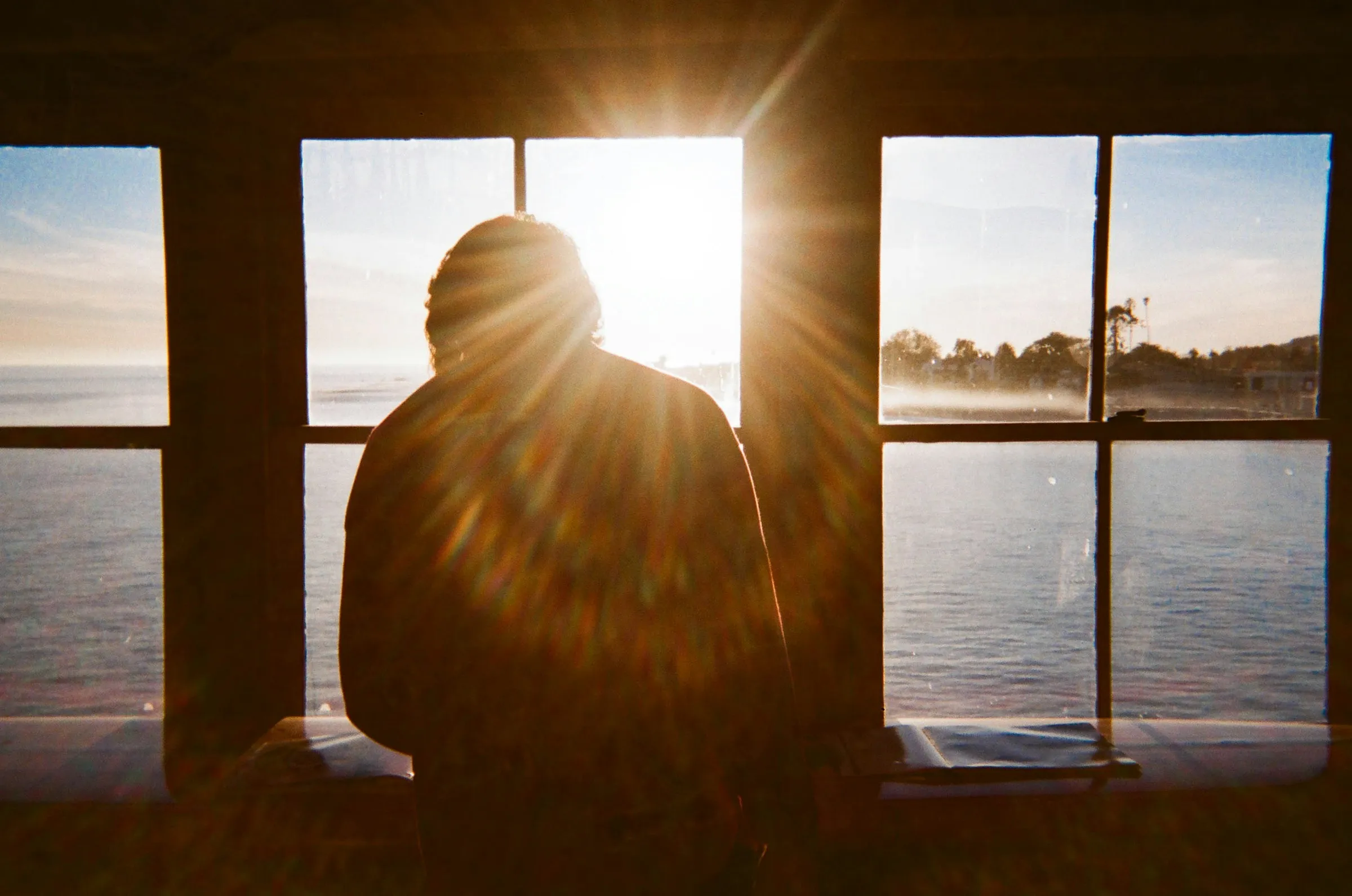 A person sitting by a sunlit window overlooking the water in the early morning