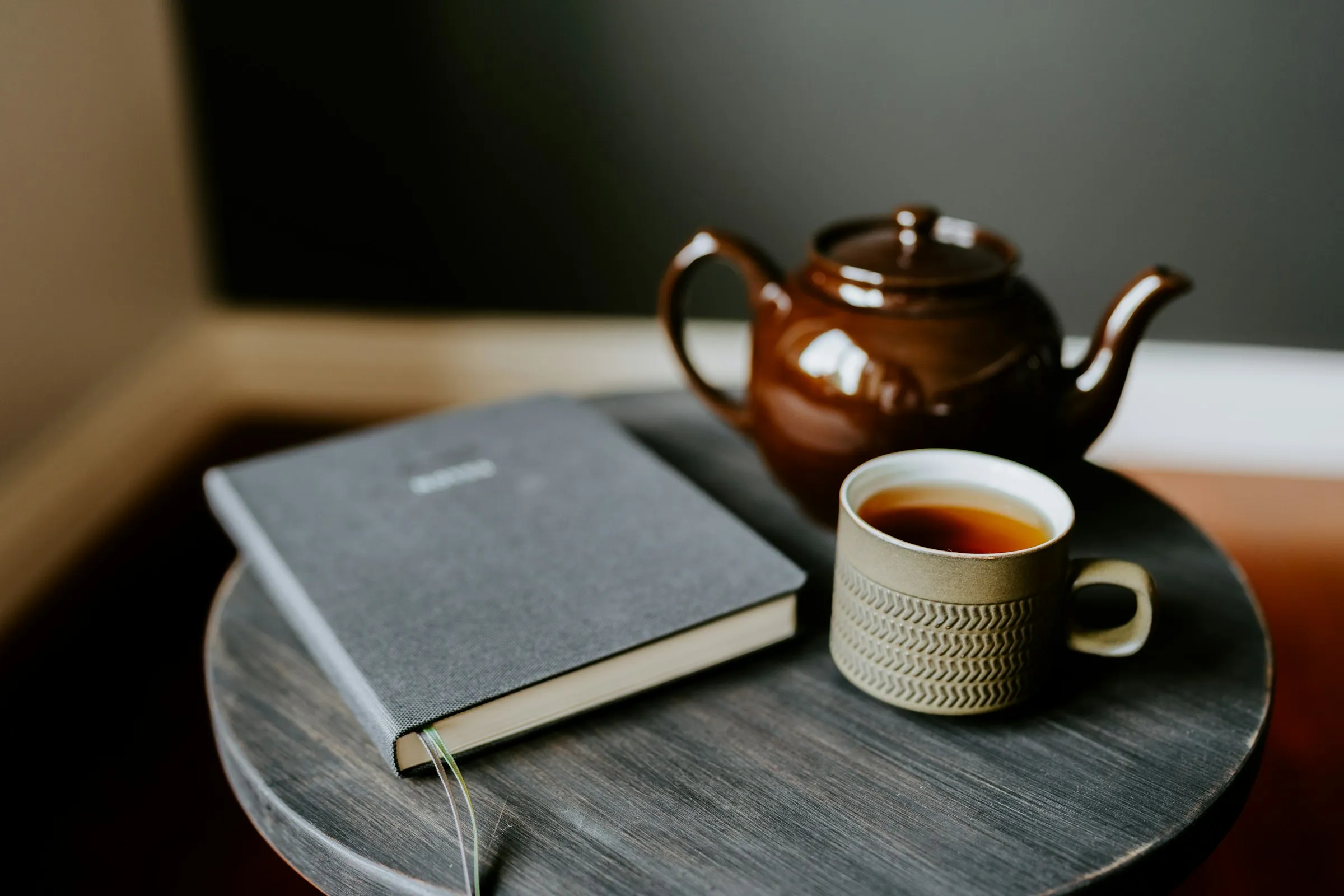 A dark notebook and a cup of tea beside a brown teapot on a small round table